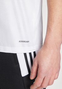 T-shirt de sport blanc avec texture en maille, arborant le logo "AEROREADY" près de l'ourlet, associé à un pantalon noir affichant des rayures blanches.