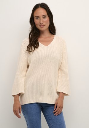 Lyse beige strikket sweater med V-udskæring, lange klokkeærmer og en afslappet pasform. Båret over blå jeans, der fremhæver en ribbet tekstur.