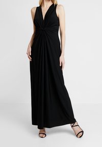 Musta, hihaton maksimekko, jossa syvä V-pääntie ja vyötäröllä solmittu kerätty yksityiskohta, valmistettu sileästä, virtaavasta kankaasta ja lattiaan ulottuvalla mallilla.