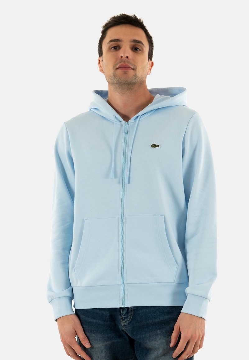 Sudadera con cremallera de color azul claro en tejido suave, que presenta un bolsillo delantero, puños de canalé y un pequeño logo verde en el pecho.