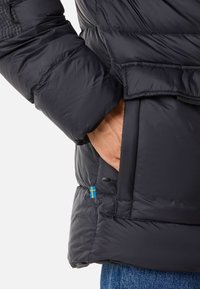 Black puffer jacket with quilted texture, featuring a side pocket and a small Swedish flag accent at the hem. Hand resting in pocket.