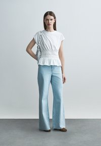 Sieviete valkā baltu blūzi ar pieguļošu vidukli un gaiši zilas plašas bikses, pār plecu nēsā sudraba krāsas somiņu, stāvot uz pelēkas grīdas.