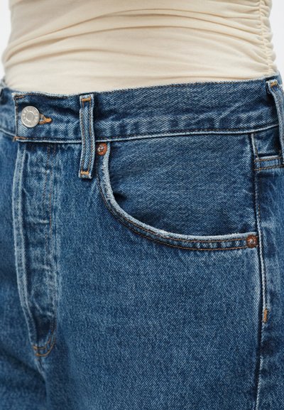 Calças de ganga azul com lavagem escura, apresentando dois bolsos frontais, rebites de cobre e um fecho clássico de botão. Detalhe do tecido texturizado visível.