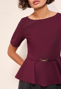 Burgundské peplum tričko s krátkými rukávy, které má přiléhavou část a ozdobnou stříbrnou sponu v pase, hladká textilní struktura.