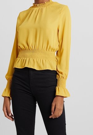 Femme portant un chemisier jaune à manches longues avec un col haut à volants, une taille et des poignets smockés, associé à un pantalon noir.