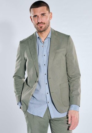 Mann steht mit den Händen in den Taschen, trägt einen hellgrünen Anzug und ein hellblaues Hemd vor einem schlichten Hintergrund.