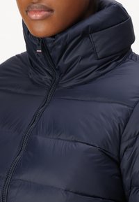 Giacca puffa blu navy con colletto alto, zip frontale e texture trapuntata. Presenta un piccolo dettaglio del logo vicino al colletto.