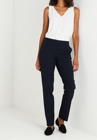 Un pantalon ajusté bleu marine associé à un top sans manches à col en V blanc avec de subtils pois argentés. Des chaussures noires à enfiler complètent le look.
