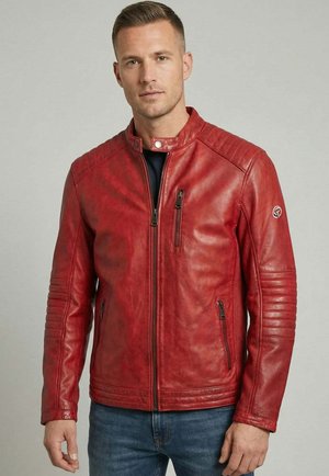 Homme portant une veste en cuir rouge cintrée avec des poches zippées, des détails matelassés aux épaules et aux manches, associée à un jean bleu sur fond gris.