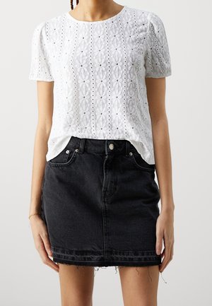 Kvinde iført en hvid kortærmet tekstureret floral top stoppet ned i et sort denimskørt med frynset kant og forlommer.