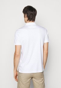 Witte polo met een lichtblauw gestippeld patroon, korte mouwen en een platte kraag, met zijsplitten aan de onderkant voor extra bewegingsvrijheid.