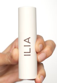 Prodotto per labbra ILIA in un contenitore cilindrico di un bianco opaco. Presenta un semplice logo marrone ed è tenuto in una mano con unghie naturali.