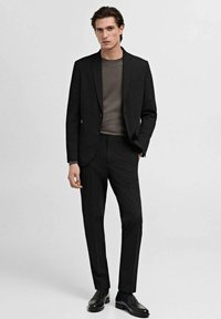 Costume noir avec veste et pantalons ajustés sur mesure, associés à un pull gris foncé. Terminé avec des chaussures en cuir noires. Texture de tissu lisse.