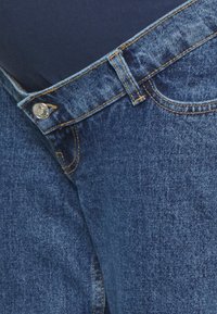 Jeans de denim azul com cintura média, costura visível e fecho em botão metálico. Apresenta bolsos frontais padrão e uma textura ligeiramente desgastada.