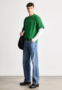 T-shirt verde de algodão sobre uma blusa de manga longa branca, combinada com jeans azuis e sapatos pretos, e uma mala de ombro preta.