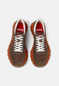 Hnědé textilní tenisky s oranžovými akcenty, bílými šňůrkami, červenými vložkami a robustní podrážkou mají výraznou texturu a masivní tvar.