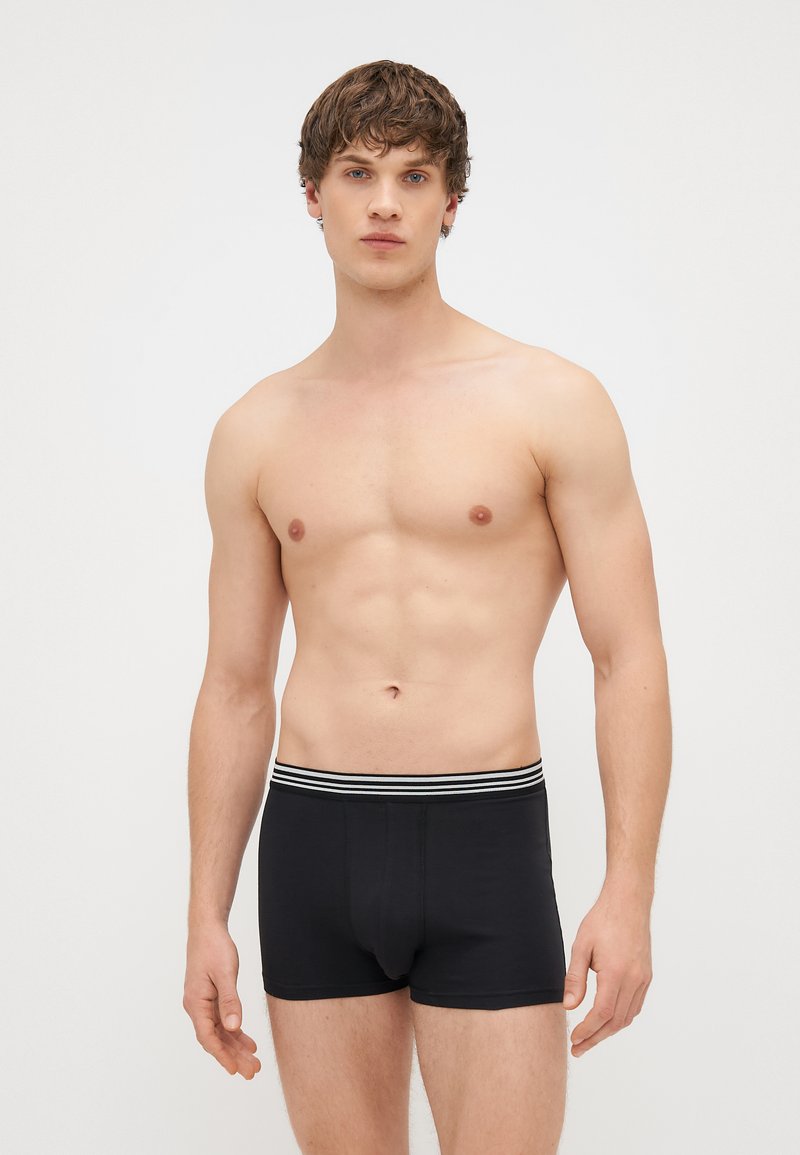 Μαύρες μποξεράκι φτιαγμένες από μαλακό ύφασμα, με ζώνη μέσης που προσφέρει άνεση και λευκές ρίγες. Στενή σχεδίαση με μέτρια ψηλή κοπή.