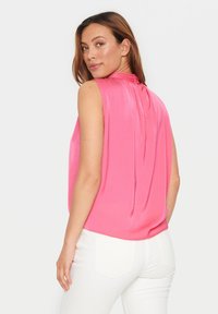 Top senza maniche e colletto alto in tessuto rosa brillante con una texture liscia. Presenta una chiusura a chiave sul retro e un design drappeggiato. Abbinato a pantaloni bianchi.