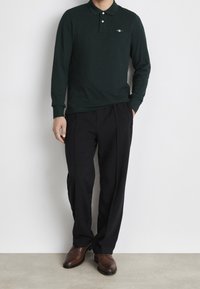 Dark green long-sleeve polo shirt with two buttons, paired with black pleated trousers and brown leather shoes, displayed against a neutral background.