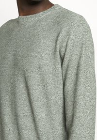 Suéter de punto verde claro con cuello redondo, que presenta una tela texturizada y detalles sutiles en todo el diseño.