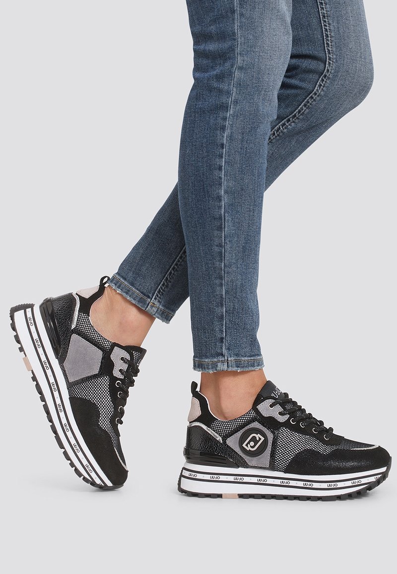 Personne portant des baskets en mesh noir et gris avec des semelles blanches et un jean skinny bleu, un pied levé derrière l'autre.