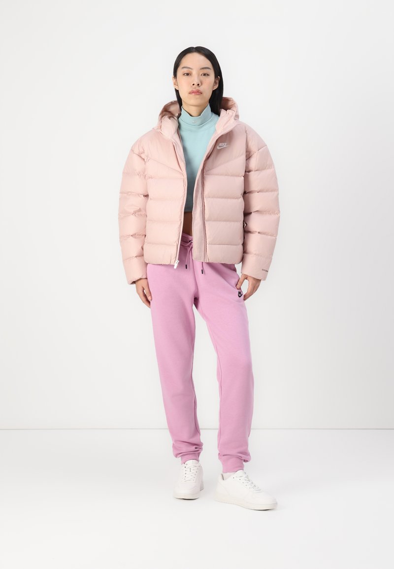 Frau, die in einer hellrosafarbenen Nike-Pufferjacke, einem mintgrünen Rollkragenpullover, rosafarbenen Nike-Sweatpants und weißen Sneakern vor einem weißen Hintergrund steht.