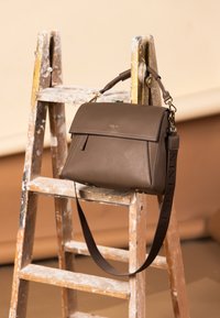 Borsa in pelle marrone con patta superiore piegata, ferrature dorate, tracolla regolabile e logo impresso, esposta su una scala di legno.