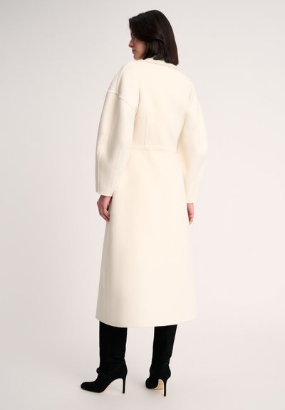 Long, cream wool coat with oversized sleeves and a structured back. Paired with black suede high-heeled shoes. Simplistic design, no patterns.