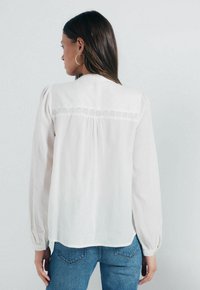 Weiße Bluse mit langen Ärmeln, strukturiertem Stoff und Spitzendetails am Schulterbereich, kombiniert mit blauen Jeans. Rückansicht dargestellt.
