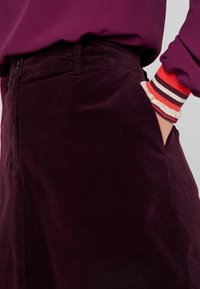 Gonna in corduroy bordeaux con tasche laterali e design a vita media, completata da un polsino a coste multicolore visibile al polso.