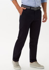 Pantaloni di cotone navy con una vestibilità tailored, dotati di tasche laterali e una cintura intrecciata grigio-nera. Abbinati a sneakers grigie chiare.