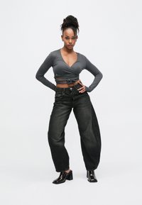 Γκρι μακρυμάνικο τοπ με δέσιμο στη μέση, συνδυασμένο με φαρδιά μαύρα τζιν και λαμπερά μαύρα παπούτσια με τακούνι.