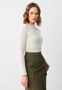 Haut en dentelle à manches longues en blanc cassé avec un motif floral et un col haut, associé à une jupe verte arrivant au genou avec des poches.