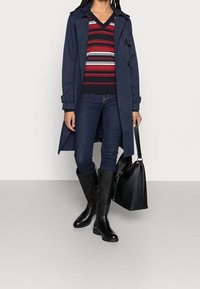 Un trench-coat navy sur un pull rayé, associé à un jeans foncé et des bottes noires montantes. Comprend un sac noir avec une bandoulière épaisse. Textures lisses.