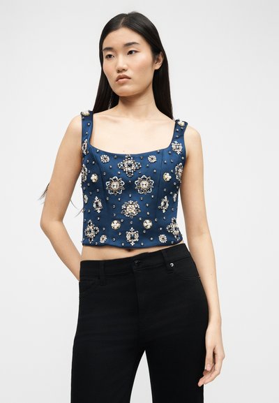 Νεαρή γυναίκα που φοράει μπλε crop top χωρίς μανίκια, διακοσμημένο με περίτεχνα ασημένια και μαργαριταρένια στολίδια, συνδυασμένο με μαύρο παντελόνι.