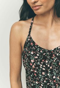 Top sin mangas negro con estampado floral que presenta pequeñas flores rosas y blancas, tirantes finos y una tela suave y ligera. Textura suave con escote en V.