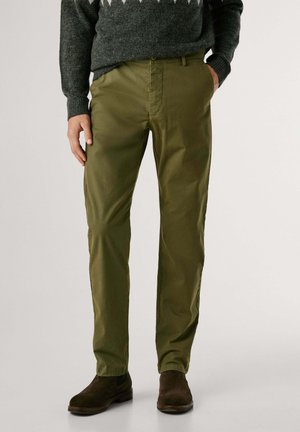 Olivgrüne Chinos mit geripptem Bund, seitlichen Taschen und leichtem Glanz. Kombiniert mit dunkelbraunen Wildleder-Chelsea-Stiefeln.