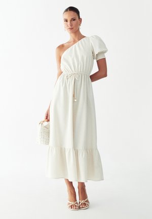 Vrouw staand, gekleed in een crèmekleurige asymmetrische jurk met een opgeblazen mouw, houdend een witte gevlochten handtas vast, en dragend crèmekleurige sandalen met riempjes.