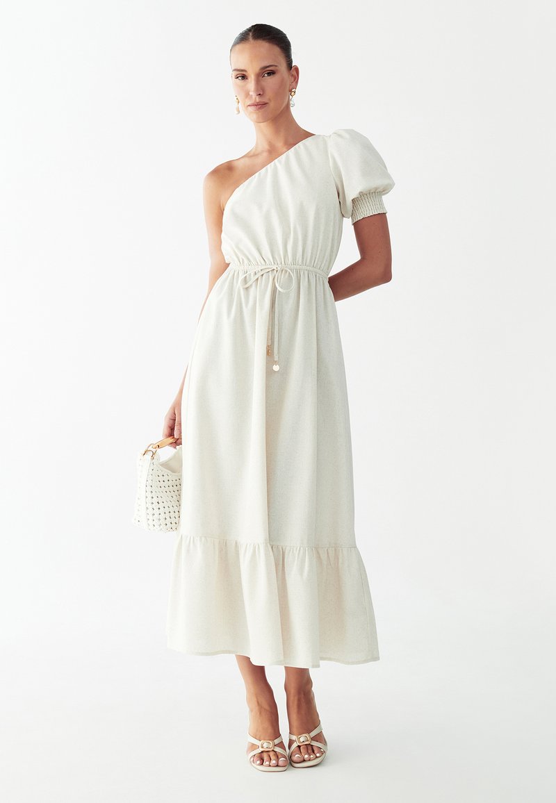 Femme debout portant une robe crème asymétrique à manche bouffante, tenant un sac à main blanc tissé, et portant des escarpins crème à lanières.