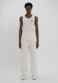 White ribbed tank top with a black logo, paired with loose-fitting white sweatpants. Model wears white athletic shoes.