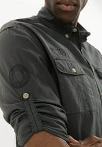 Chemise boutonnée gris foncé avec des manches retroussées, des poches poitrine et des détails brodés sur les manches. Fabrication en tissu doux et léger.