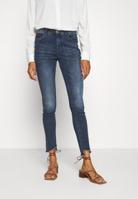 Hubené džíny v tmavě modré barvě s vybledlým vzhledem, zakončené surovým lemem. Spárované s světlou blůzou a hnědými sandály na vysokém podpatku.