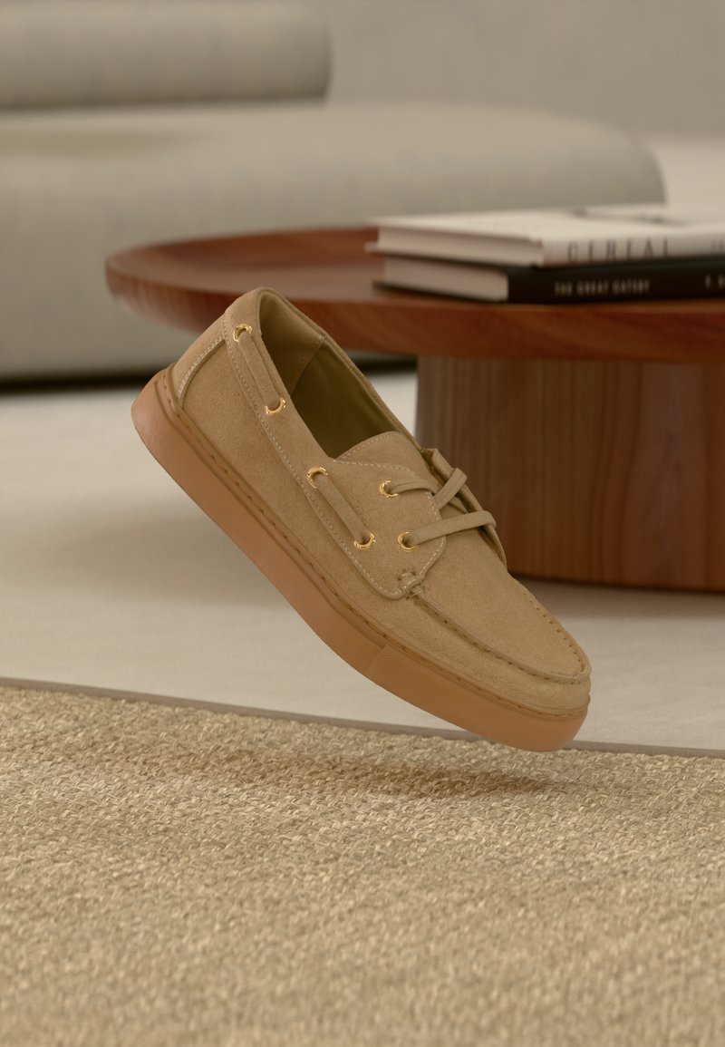 Sapato casual bege em camurça com sola e atacadores castanho claro, colocado sobre um tapete bege texturizado em frente a uma mesa de café de madeira com livros empilhados.