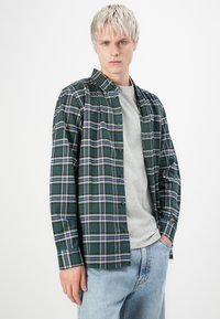 Groene geblokte shirt met blauwe en gele accenten, met knoopsluiting, lange mouwen, gedragen over een grijze T-shirt, gecombineerd met lichtblauwe jeans.