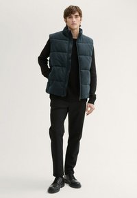 Schwarze Pufferweste mit hohem Kragen, gesteppter Design, dunkelgrüne Farbe, kombiniert mit einem schwarzen strukturierten Pullover und schmal geschnittenen Hosen, schwarze Schuhe.
