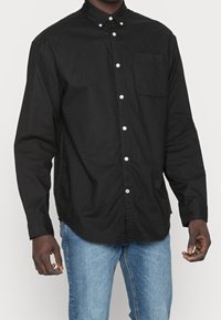 Camicia nera button-up in cotone, con colletto, taschino frontale e bottoni bianchi, abbinata a jeans azzurri chiari.