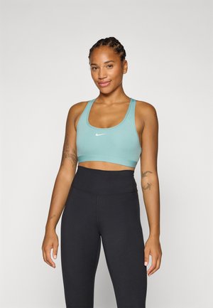Smiling woman wearing a light blue sports bra and black leggings, standing confidently against a plain white background.