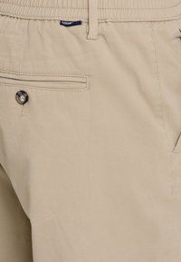 Beżowe spodnie z bawełny z dodatkiem innych włókien, wyposażone w kieszeń z przodu z zapięciem na guzik oraz płaskim kroju. Gładka struktura z minimalnymi detalami przeszycia.