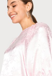 Top de cetim rosa com padrão floral em relevo, mangas longas e decote redondo, apresentando uma textura suave e um brilho sutil.