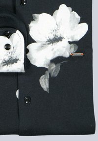 Schwarzes Stoffhemd mit weißem Blumenmuster, schwarzen Knöpfen und einem markanten Kragen. Enthält ein Etikett mit der Aufschrift "emiloadan."